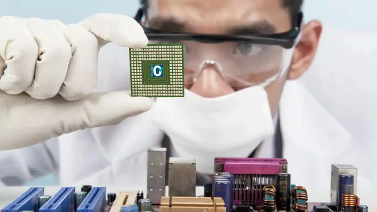 Technician in cleanroom gear holding a microchip over a circuit board